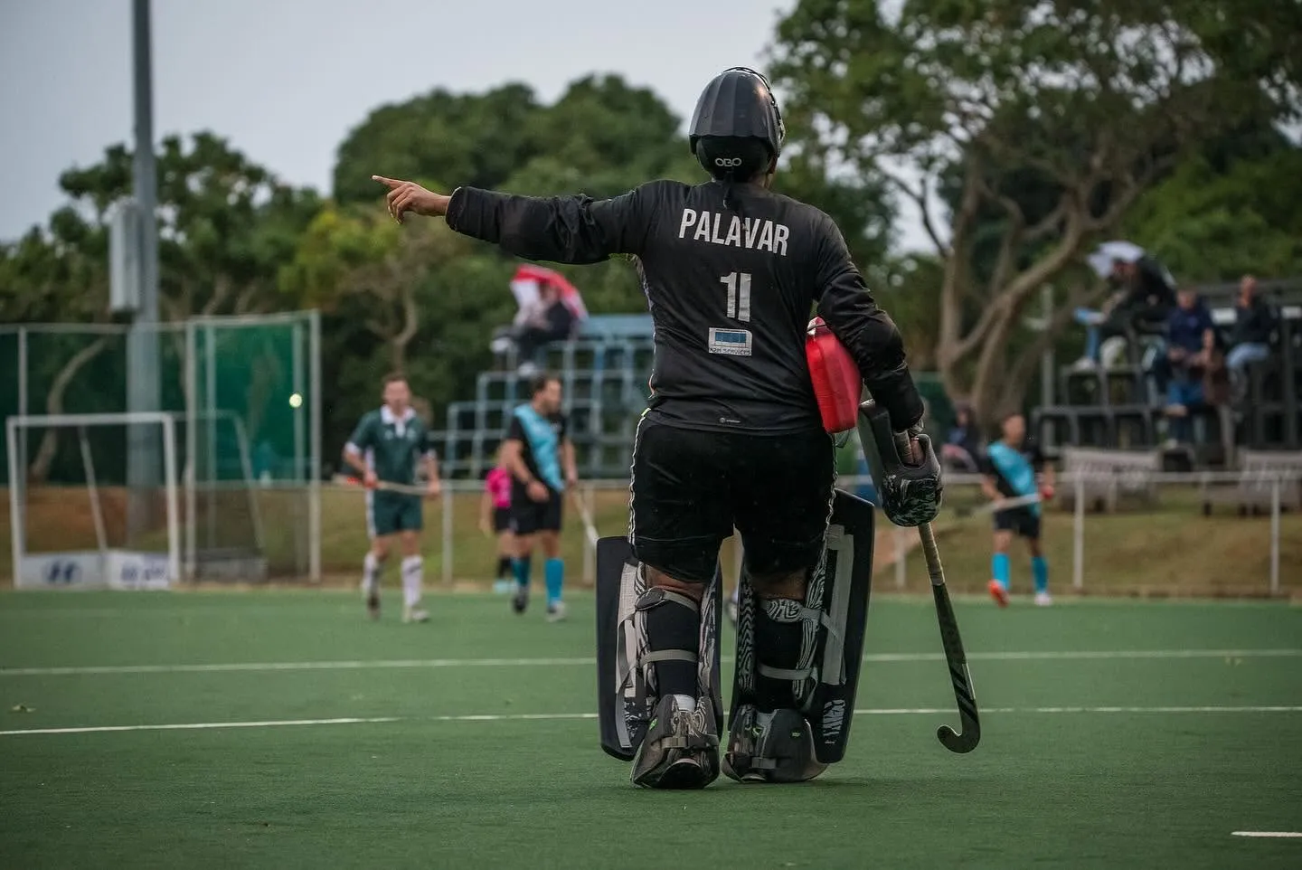 A goalkeeper in full kit points upfield during an Impis men's match, with players and spectators behind.
