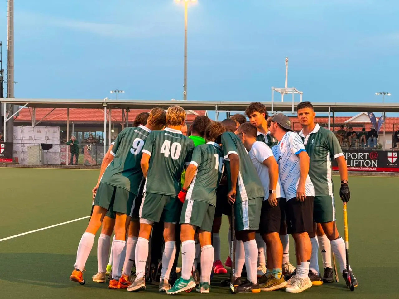 Impis men's players and coaches huddle together on the turf under the floodlights at dusk.